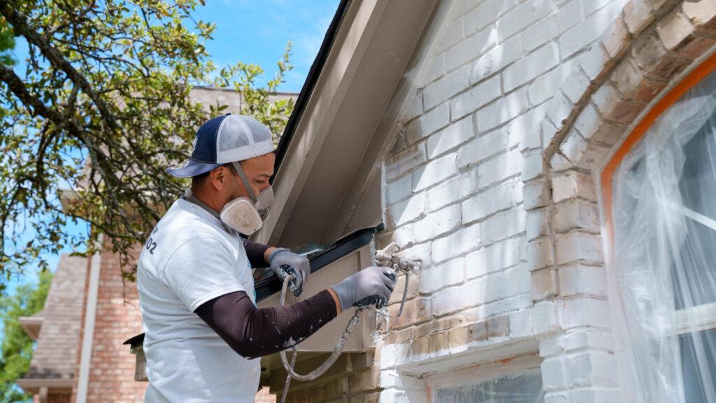 Professional painter using a spraying tool to apply white paint to a tan brick home_That 1 Painter Arlington Alexandria