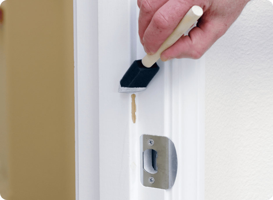 Hand using a foam brush to touch up chipped paint on a door frame for repair services with that 1 painter