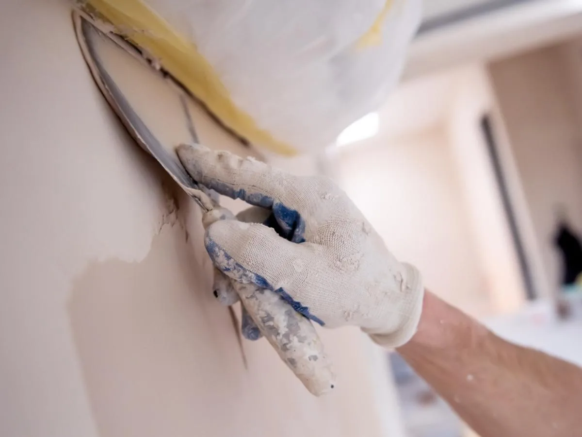 Gloved hand smoothing joint compound over a drywall patch with a putty knife before painting