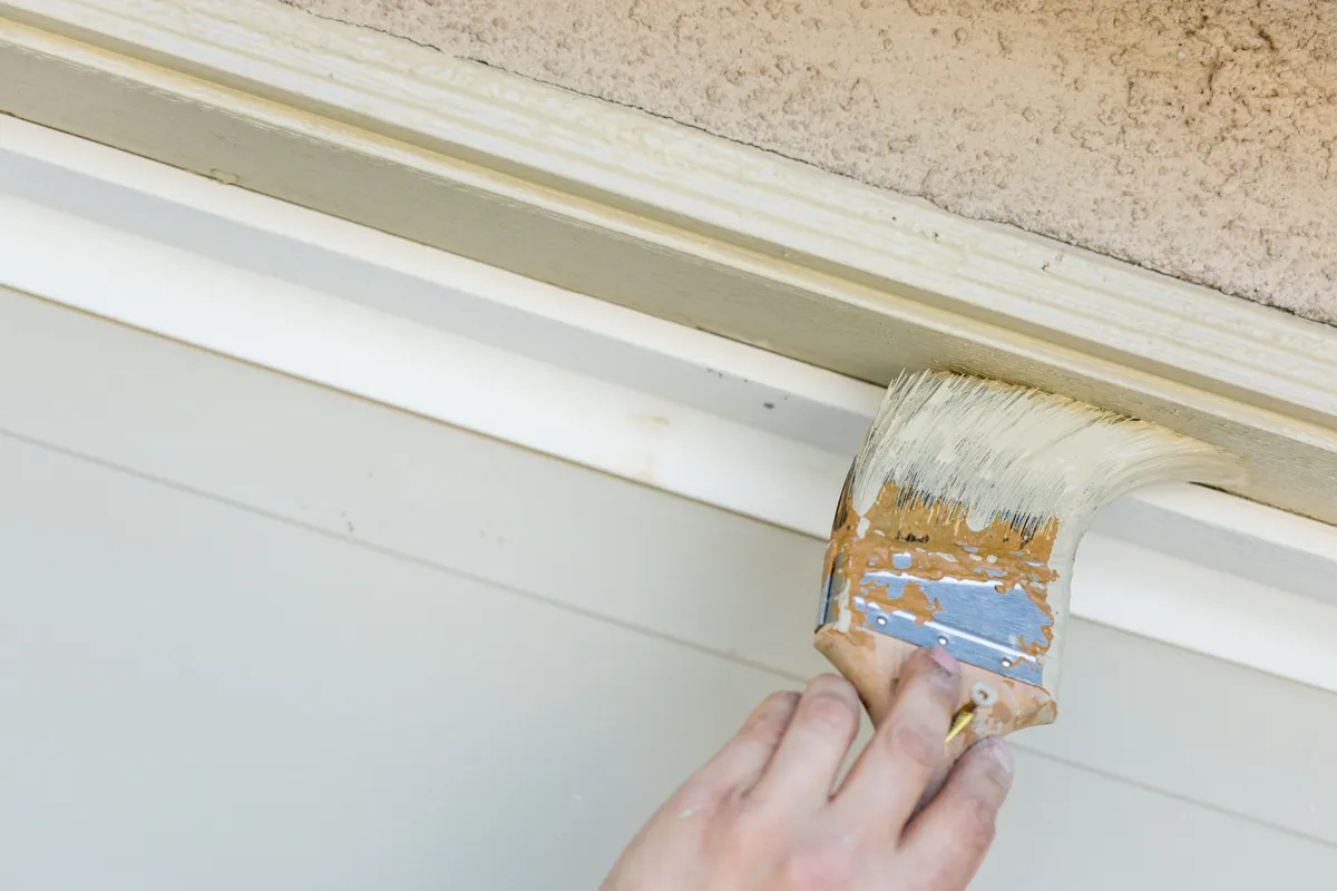 Paintbrush applying white paint to crown molding trim