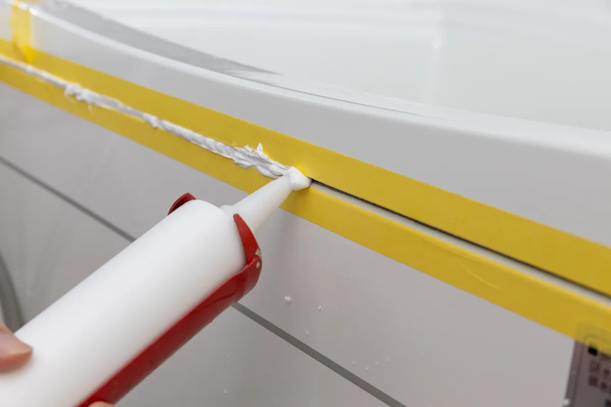 Caulk gun applying a fresh bead along a bathtub with yellow painter's tape