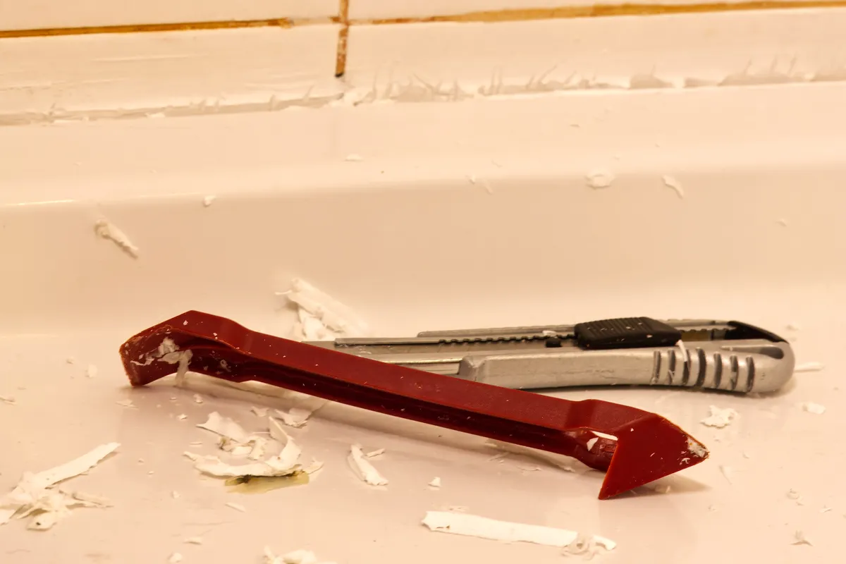 Red scraper and utility knife beside strips of removed caulk on a bathtub ledge