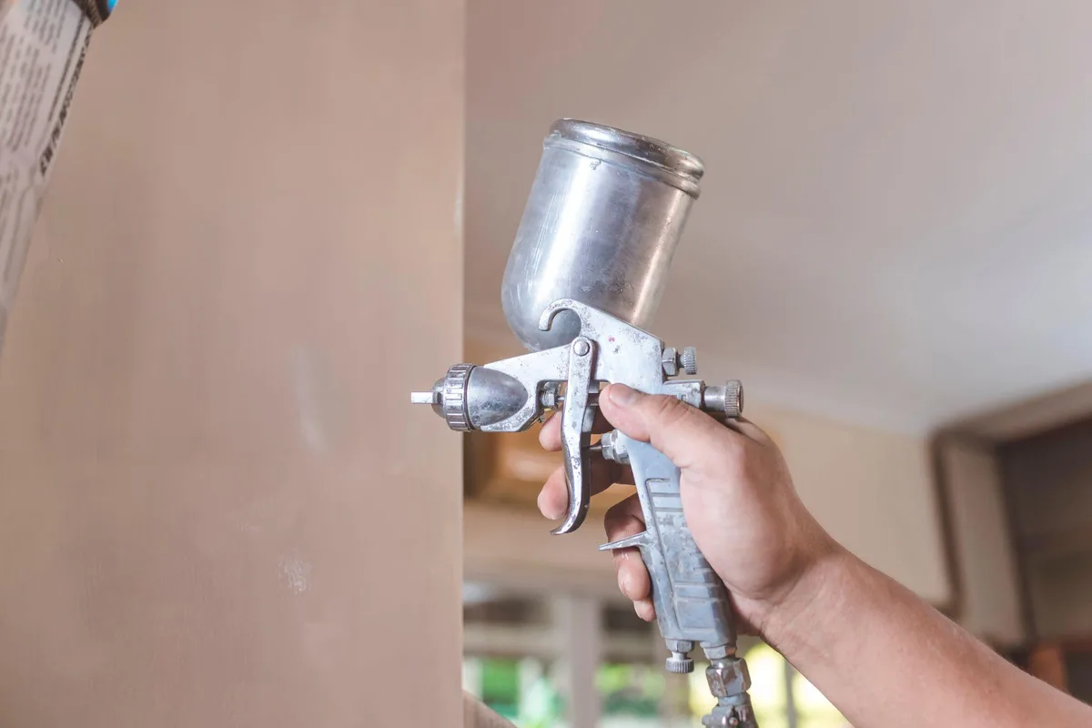 Hand holding a gravity-feed spray gun beside a freshly painted beige wall