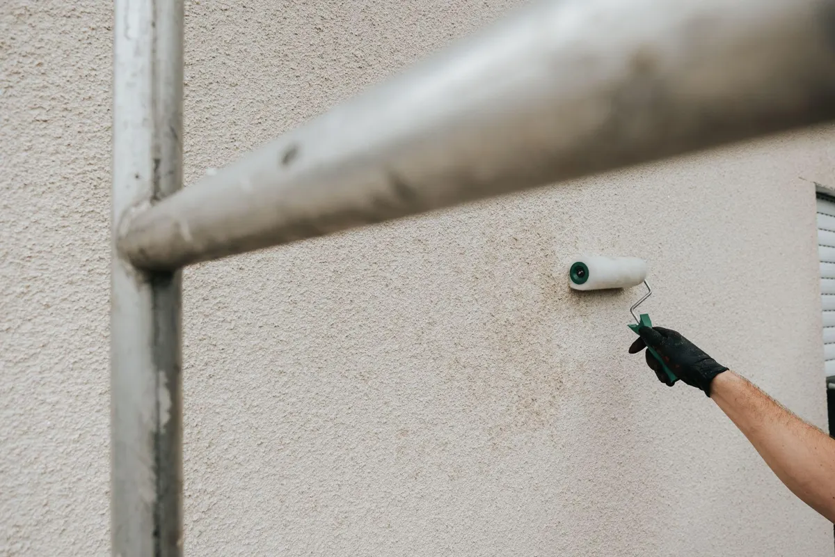 Gloved hand rolling paint onto a textured exterior stucco wall beside scaffolding