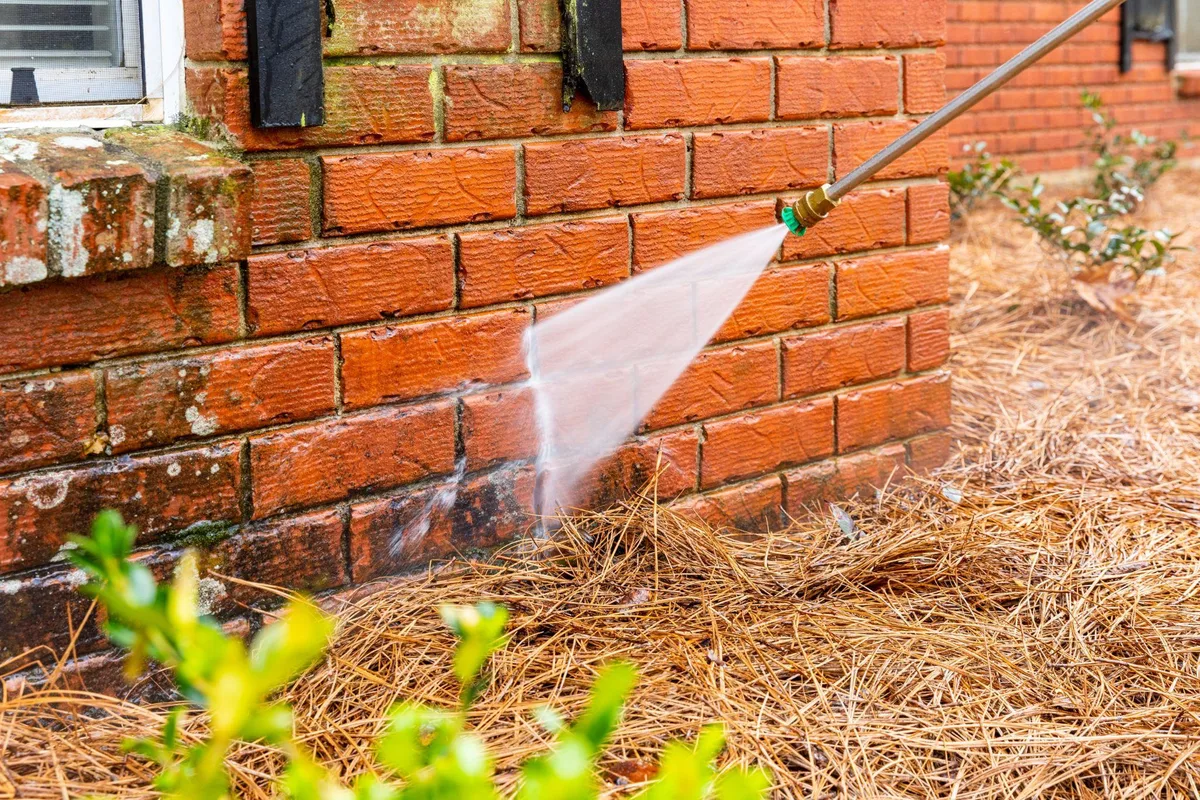 Pressure washer wand spraying water at the base of a red brick house wall above pine straw
