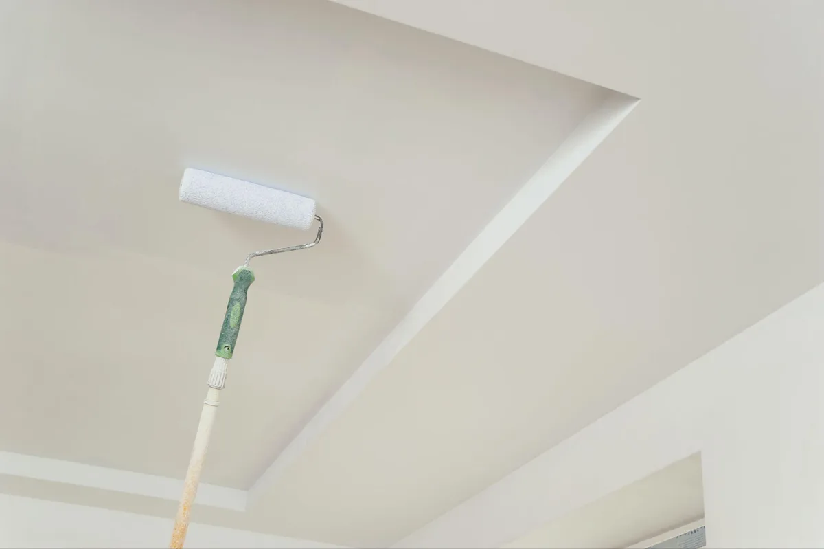 Paint roller on an extension pole rolling white paint onto a recessed tray ceiling