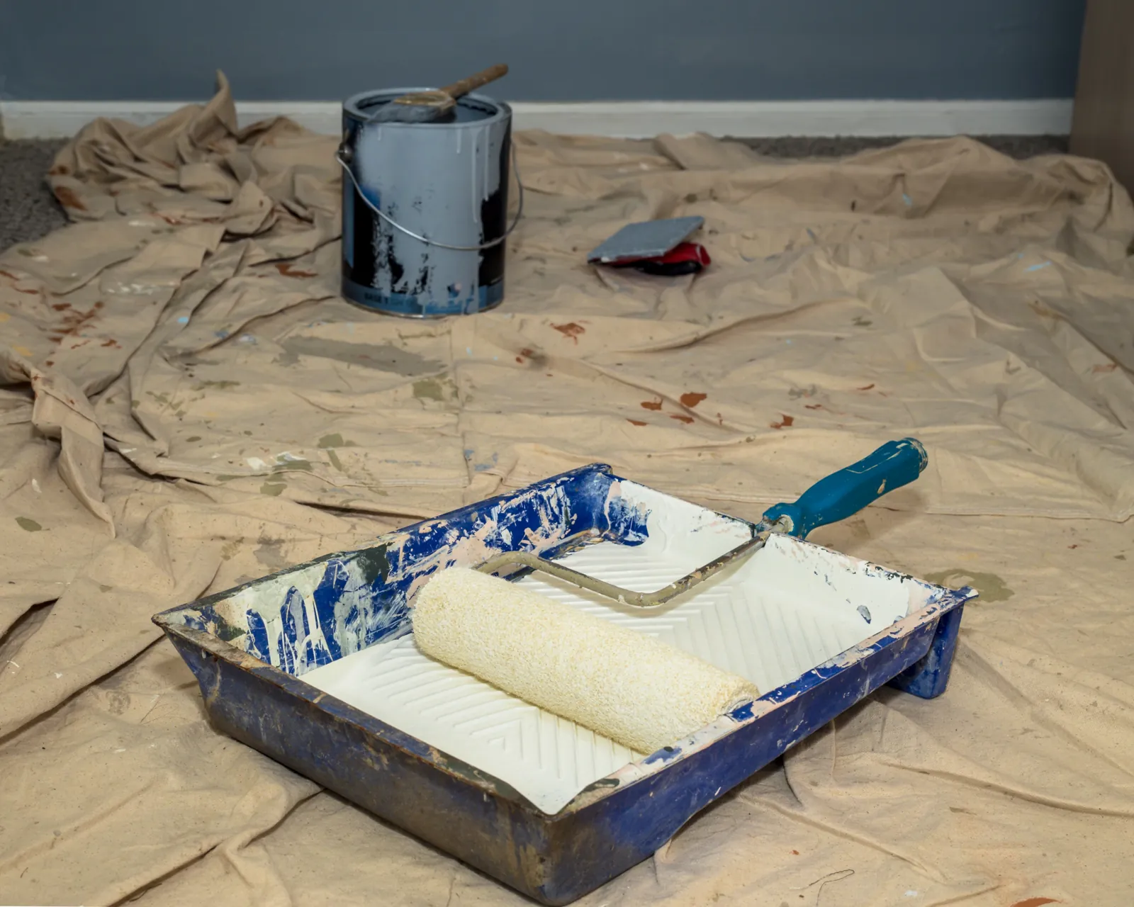 Paint roller resting in a tray of white paint on a drop cloth beside an open paint can
