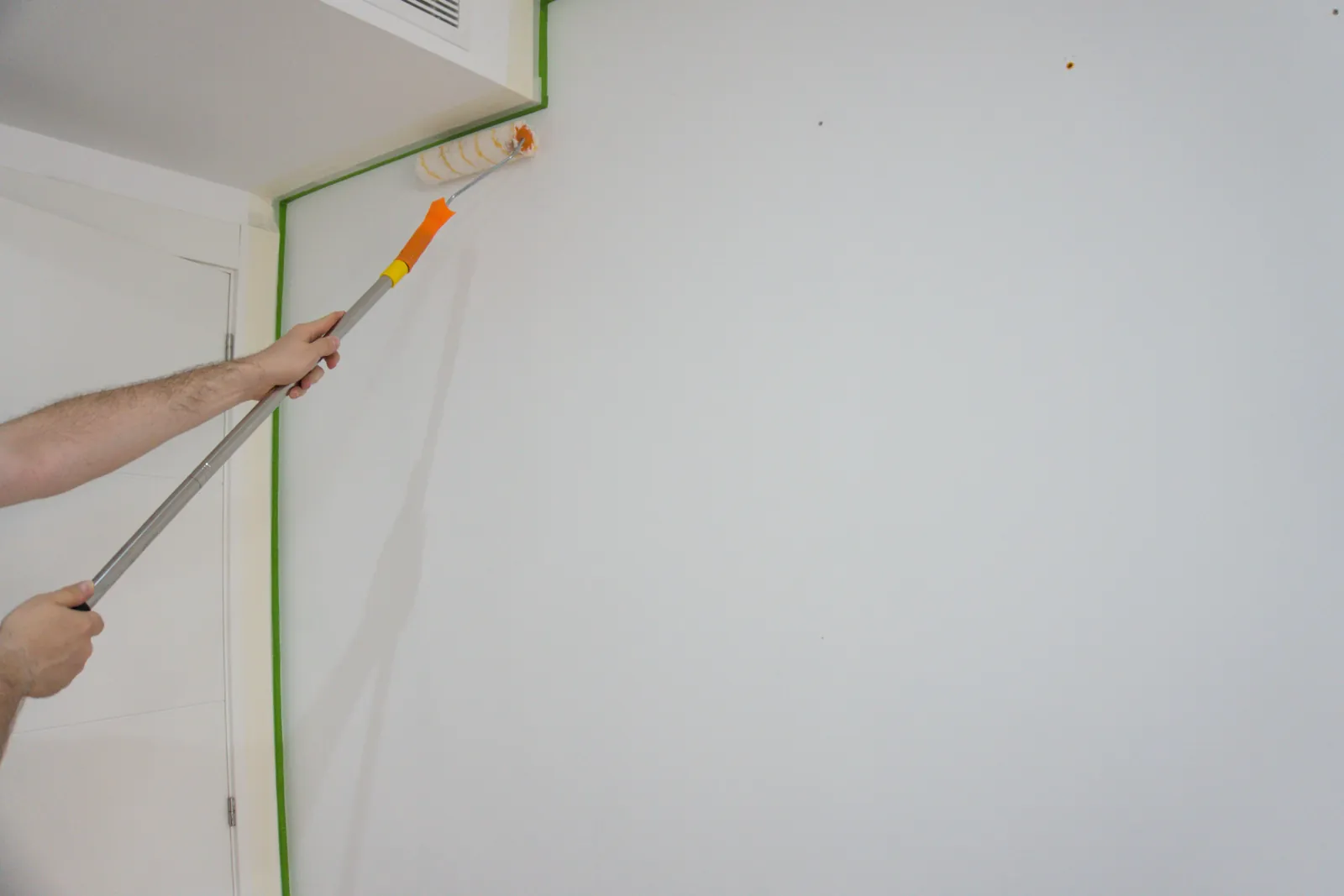 Painter using a roller on an extension pole beside green painter's tape on a white wall