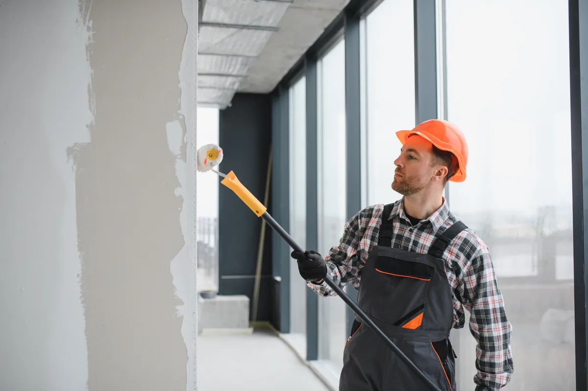 Painter in an orange hard hat rolling primer onto a wall with an extension pole