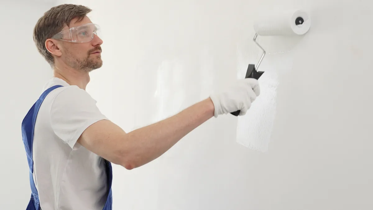 Painter in blue overalls and safety glasses rolling white paint onto a wall