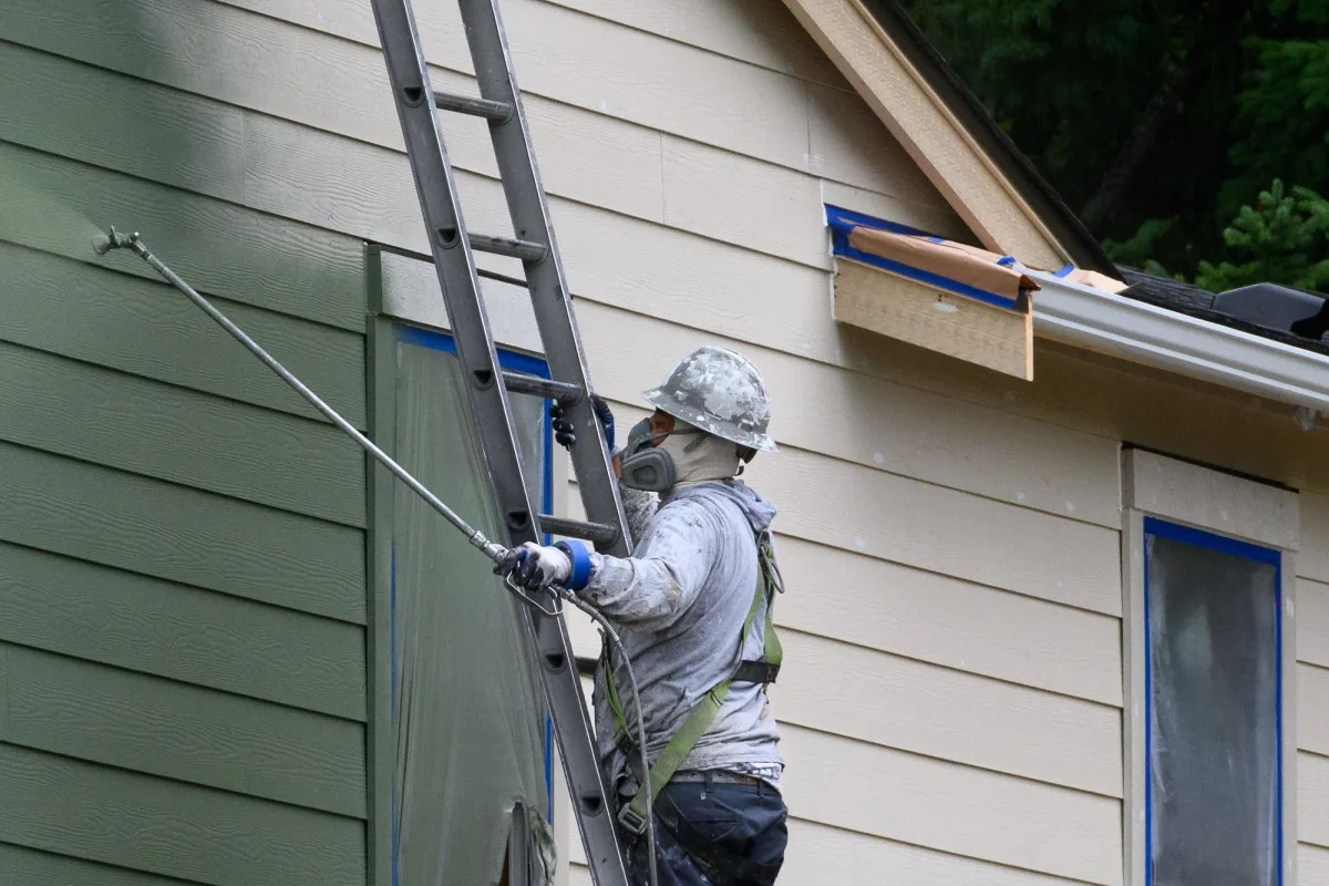 Painter on a ladder spraying dark green paint onto lap siding with the windows masked off
