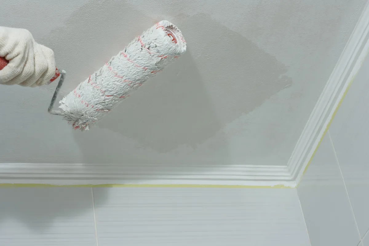 Roller painting a bathroom ceiling white with masking tape along the tile