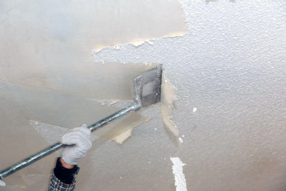 Gloved hand pushing a pole scraper across a popcorn ceiling, with scraped and textured sections side by side