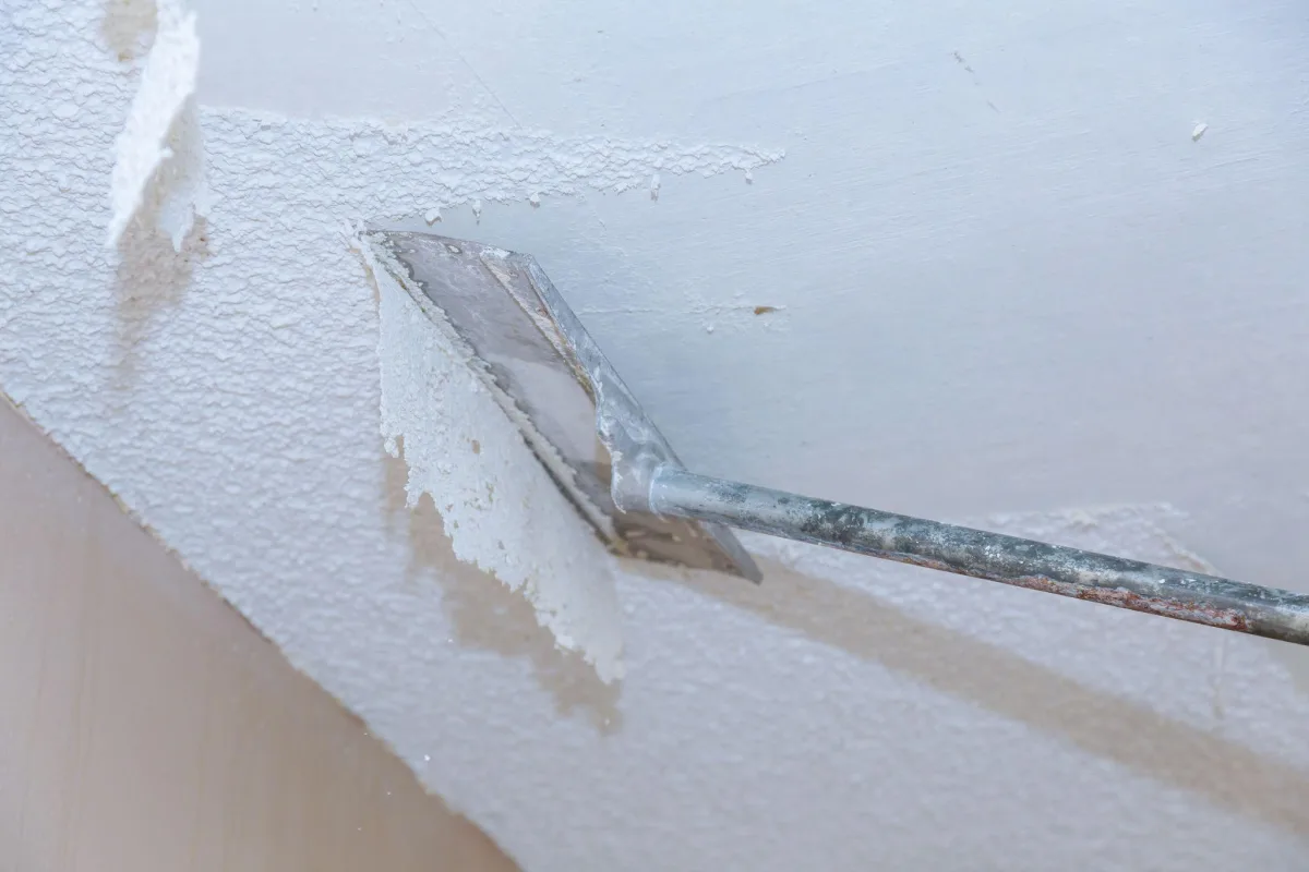 Long-handled scraper peeling wet popcorn texture off a ceiling to reveal smooth drywall