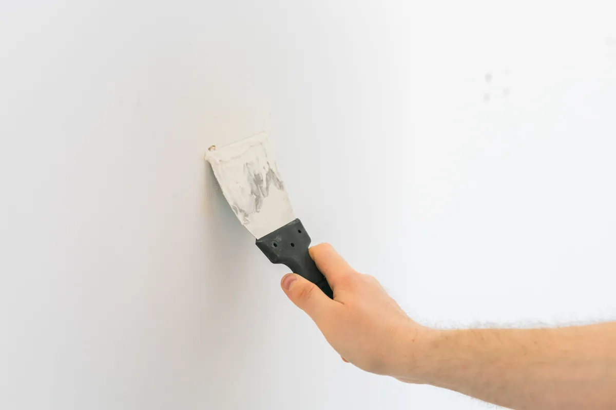 Hand pressing a putty knife against a white wall to fill a small hole
