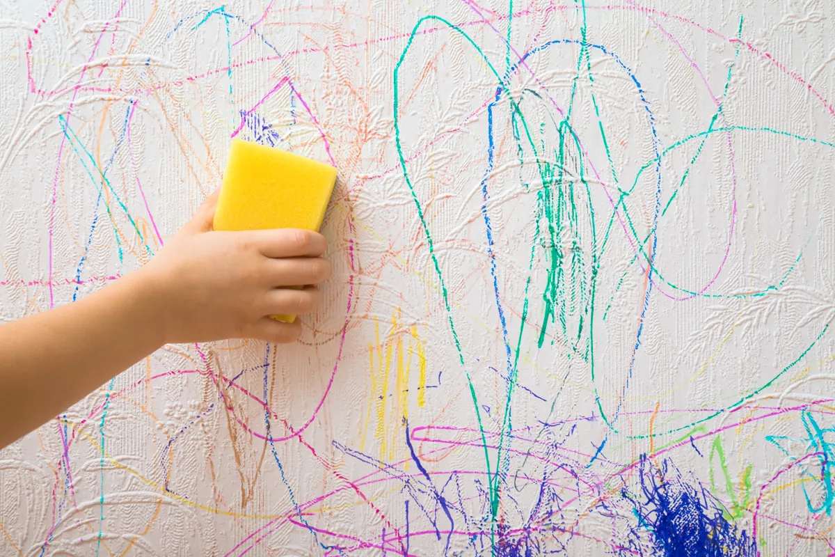 Child's hand using a yellow sponge to wipe colorful crayon marks off a white wall