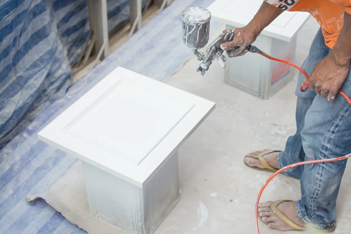 Worker spray painting a white cabinet door with a gravity-feed spray gun