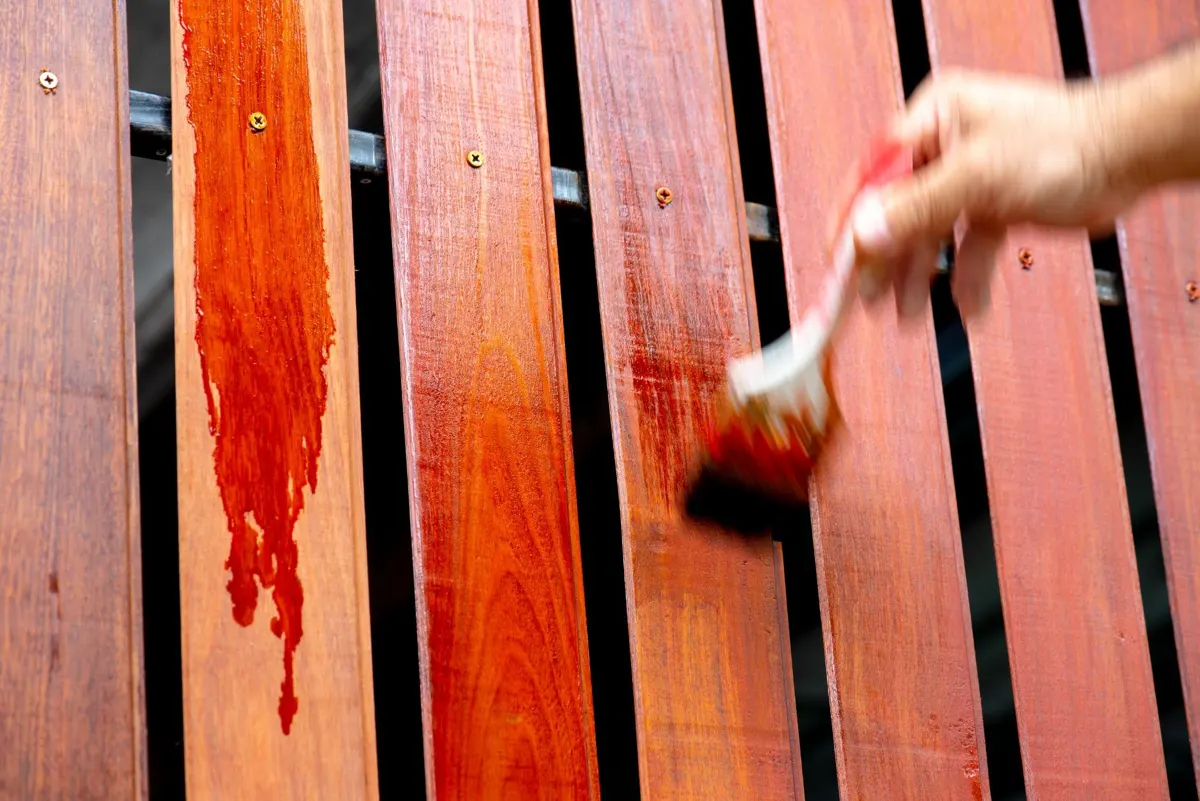 Hand brushing reddish stain onto vertical wood fence boards, with a drip running down one board