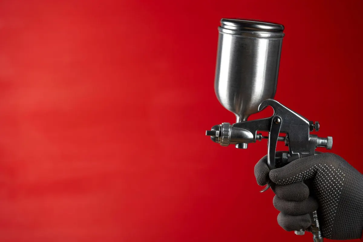 Gloved hand holding a gravity-feed spray gun against a red background