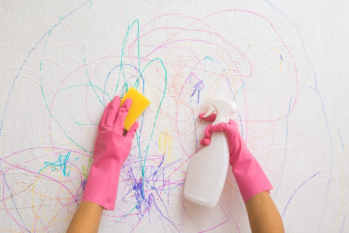 Hands using a sponge and spray cleaner to wash colorful marker scribbles off a white wall