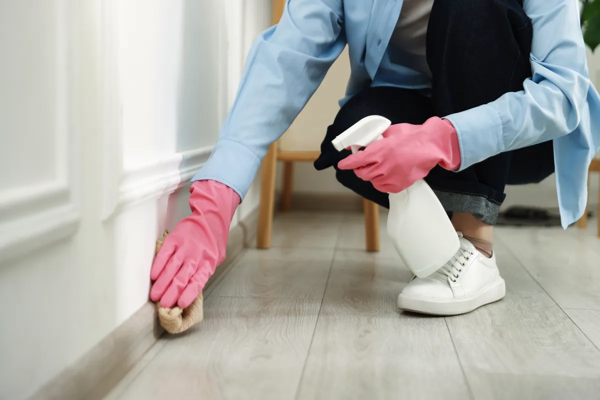 Woman in pink gloves cleaning a baseboard with a spray bottle and sponge