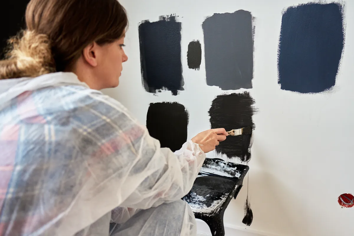 Woman brushing dark gray and black paint samples onto a white wall