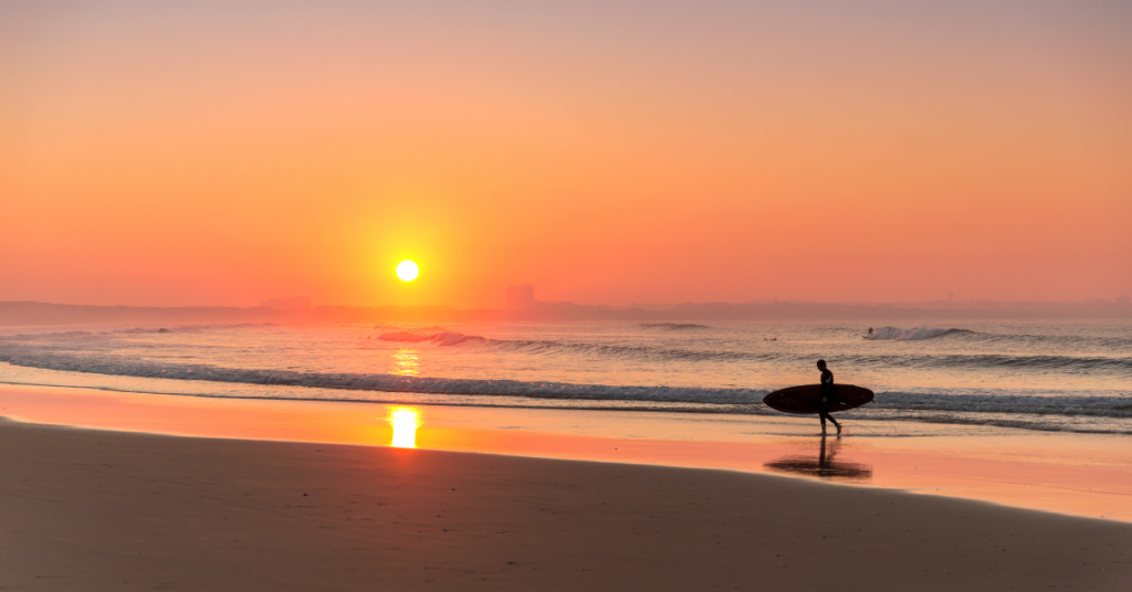 An image of a Florida sunset and a surfer walking along the beach with his board.