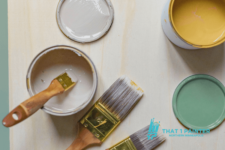 Close-up of paintbrushes and open cans of beige, green, and yellow paint, ideal for cabinet and interior painting projects. This image represents the premium materials and tools used by That 1 Painter, trusted for professional cabinet painting services in North Minneapolis, ensuring high-quality and durable finishes.
