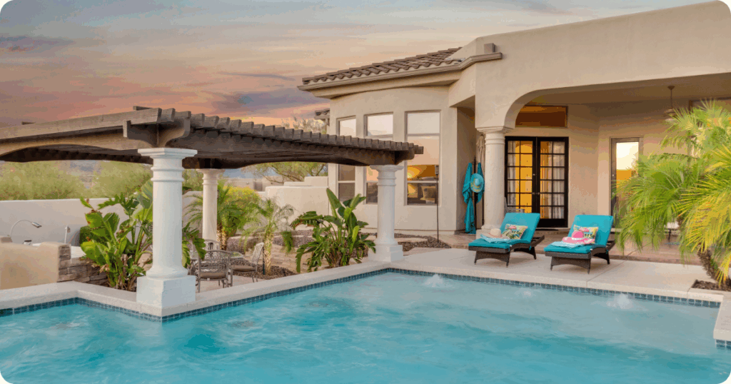 Exterior painting in Scottsdale, AZ of a luxury stucco home with a tile roof, covered patio, and poolside pergola surrounded by tropical landscaping at sunset by that 1 painter.