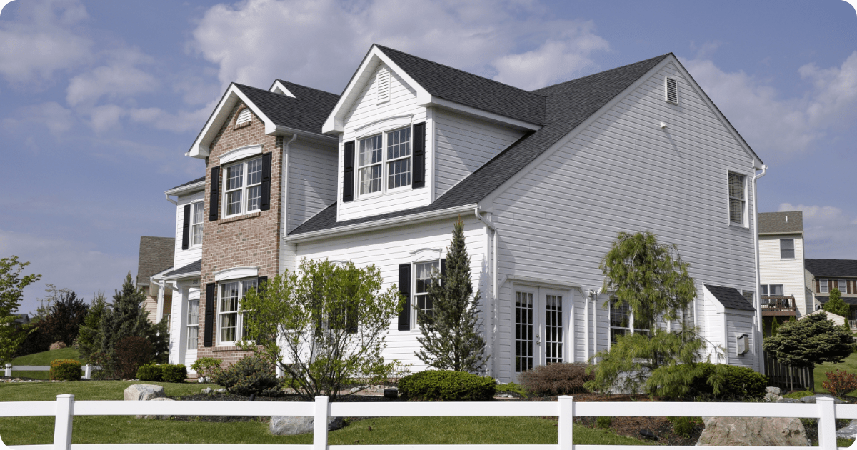 Exterior painting in Scranton of a two-story suburban home featuring white vinyl siding, red brick accent facade, and black shutters, framed by a white picket fence by that 1 painter.