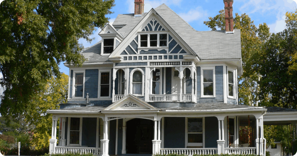 Exterior painting in Shreveport of a grand Queen Anne Victorian home with intricate woodwork, blue siding, white trim, and dual red brick chimneys by that 1 painter.