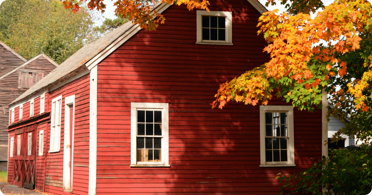 Exterior painting in Southern New Hampshire of a historic red barn-style building with white trim and aged clapboard siding beneath colorful fall foliage by that 1 painter.