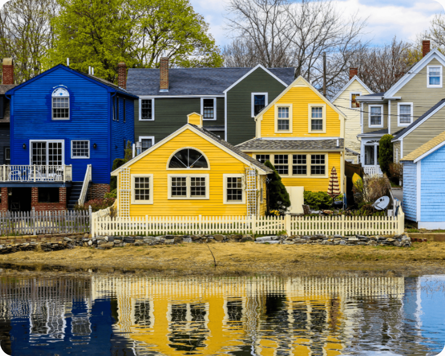 Exterior painting in Southern New Hampshire of vibrant coastal homes in shades of blue, yellow, and green with white picket fences reflecting on waterfront by that 1 painter.