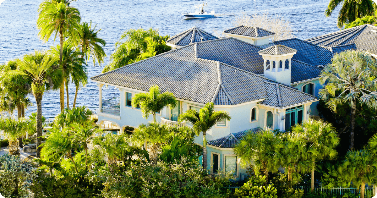 Exterior painting in Tampa Bay of a waterfront two-story home with white stucco exterior, decorative balustrades, and a spacious patio lined with palm trees by that 1 painter.