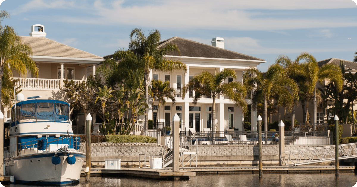 Exterior painting in Tampa Bay of a luxurious coastal home with light pastel stucco, barrel tile roofing, multiple rooflines, and dense tropical landscaping overlooking the water by that 1 painter.