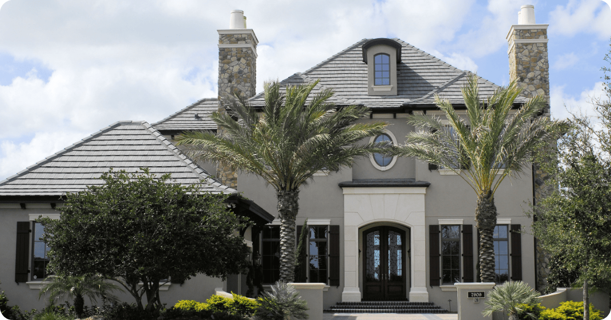 Exterior painting in Tampa Bay of an elegant French Provincial-style residence with neutral taupe stucco, stone chimney accents, dark shutters, and manicured landscaping by that 1 painter.