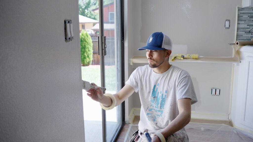 A man using a regular paint brush to carefully repaint the exterior wall of a house, giving it a fresh, renewed look under natural daylight.