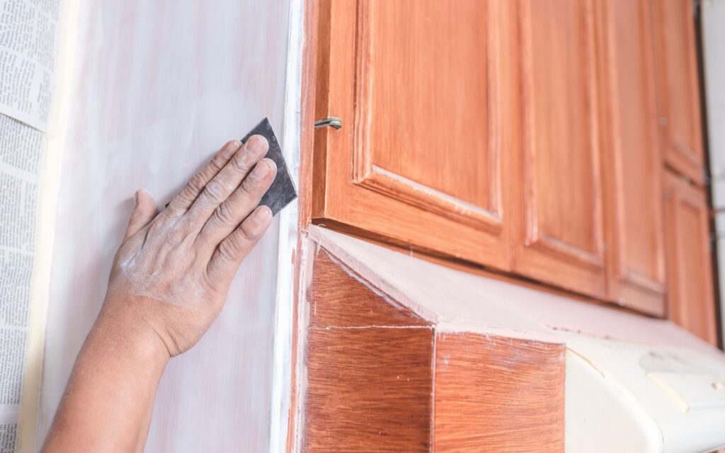What Paint Is Best for Cabinets? 2 A hand with sandpaper prepping the side of wood cabinetry for cabinet painting